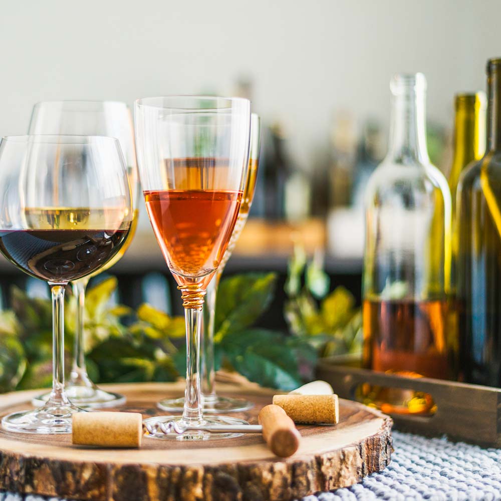 Four wine glasses containing different types and colours of wine, on a round board with a live bark edge. Corks are strewn around the base of the board. Wine bottles are visible in the background.