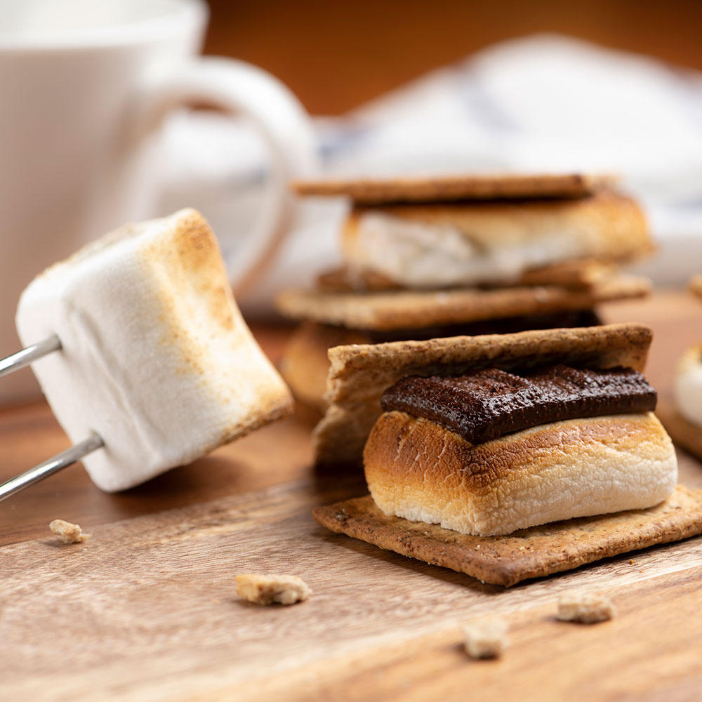 A toasted marshmallow on a skewer sits on a wooden surface beside two graham-wafer s'mores with marshmallow and chocolate. ©istockphoto by kuppa_rock