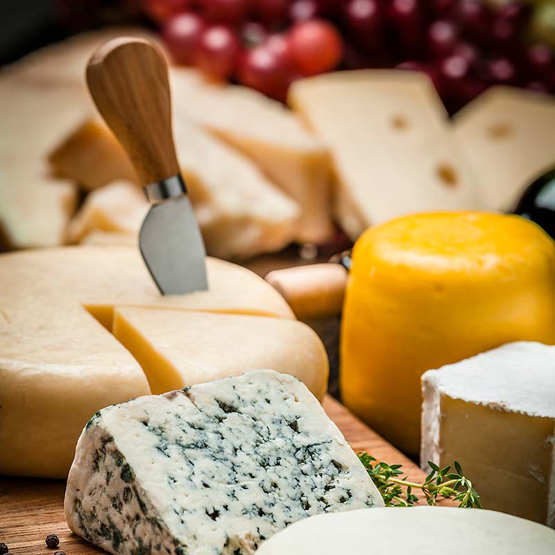 A cheese knife is embedded in a wheel of cheese. A selection of other cheeses sit on a wooden cutting board. ©istockphoto by fcafotodigital