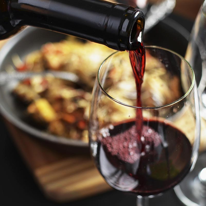 Pouring a glass of red wine into a wine glass. A plate of food is in soft focus in the background. Photo by pau_noia0 via pixabay.com.